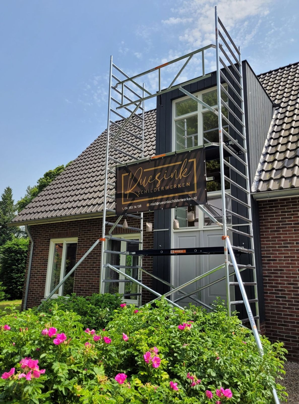 Steiger tegen een woning met het spandoek van Ruesink Schilderwerken, rozen op de voorgrond